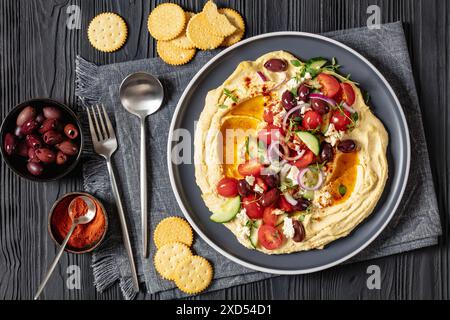 Griechischer Hummus mit frischen Gurken, Oliven, Tomaten, Fetakäse, roten Zwiebeln und Thymian auf einem schwarzen Holztisch mit Crackern, Löffel und Stockfoto