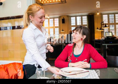 Modische gekleidete Frau zum Mittagessen modische gekleidete Frau zum Mittagessen, freundlich serviert von einer jungen erwachsenen Kellnerin. Tilburg, Niederlande. MRYES Tilburg Cafe Dudok Noord-Brabant Nederland Copyright: XGuidoxKoppesxPhotox Stockfoto