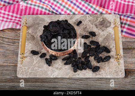 Schwarze Rosinen mit Samen. Getrocknete schwarze Rosinen in einer Kupferschale serviert auf einem Marmortablett, Rosinen verstreut um eine Kupferschale Rosinen. Stockfoto
