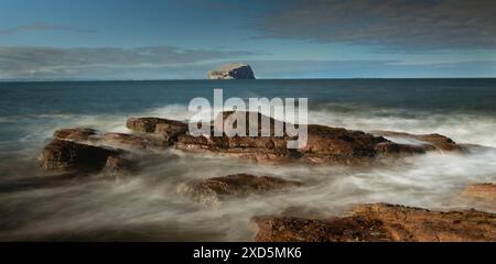 Horizontaler Blick auf Bass Rock an der Ostküste Schottlands in der Nähe von North Berwick, von einem felsigen Ufer aus, an dem Wellen auf den Felsen krachen. Lange Belichtung Stockfoto