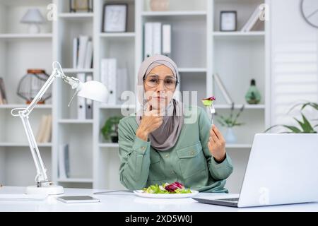 Geschäftsfrau, die einen Hijab trägt, isst einen gesunden Salat während des Mittagessens in ihrem modernen Büro mit einem Laptop und einer Lampe auf ihrem Schreibtisch. Stockfoto