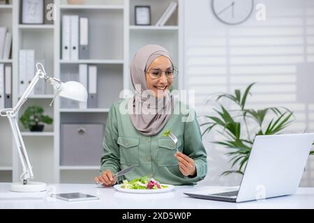 Lächelnde Frau, die Hijab trägt und gesunden Salat isst, während sie an einem Laptop in einem modernen Büro arbeitet. Konzept von gesunder Ernährung, Produktivität und positivem Arbeitsumfeld. Stockfoto