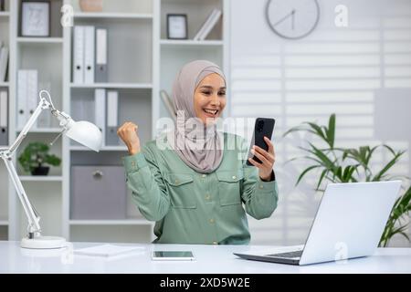 Glückliche Frau im Hijab, die Erfolg feiert, während sie im modernen Home-Office auf das Smartphone blickt und Aufregung und positive Energie zeigt. Stockfoto