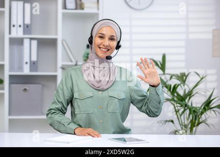 Glückliche Frau mit Headset und Hijab, die während eines Videoanrufs winkt. Sie sitzt an einem Schreibtisch in einer hellen, modernen Büroumgebung. Stockfoto