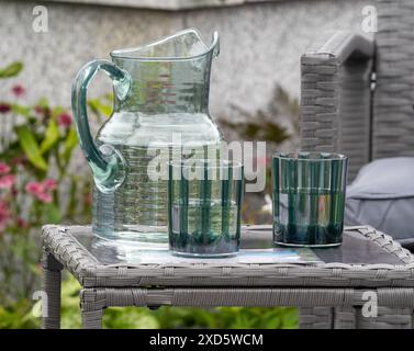 Glaskrug und zwei Trinkgläser auf einem grauen Rattan-Gartentisch. Stockfoto