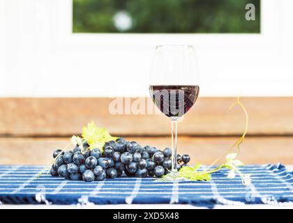 Ein Glas Rotwein und ein paar Trauben auf einem rustikalen karierten blauen Tisch in der Nähe des Sommerhäuschen Gartens. Roter Naturwein, Trauben Stockfoto