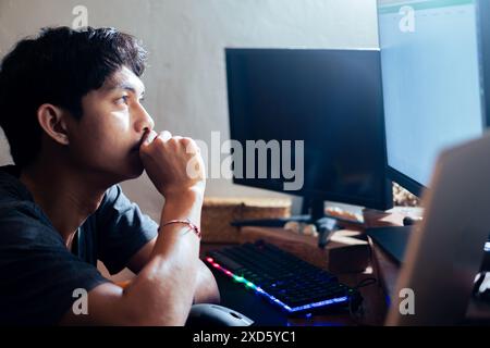 Ein junger Mann, der an einem Schreibtisch sitzt und auf einen Computerbildschirm blickt. Er scheint tief in Gedanken zu sein, mit der Hand auf dem Kinn. Der Schreibtisch ha Stockfoto