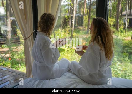 Zwei Frauen in weißen Bademänteln schlürfen Tee, während sie an einem Fenster mit Blick auf den Wald sitzen und einen entspannenden Moment genießen. Freunde entspannen in einem Landhaus in der Natur. Stockfoto