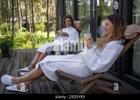 Zwei Frauen in weißen Bademänteln liegen auf Deck mit Kaffee, umgeben von Wald, und genießen einen friedlichen Rückzugsort. Freunde trinken ein duftendes erfrischendes Getränk zum Frühstück, bevor sie einen wunderbaren Tag voller Outdoor-Aktivitäten genießen. Stockfoto