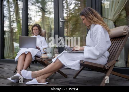 Zwei Frauen in weißen Bademänteln, die Laptops benutzen, während sie auf einer von der Natur umgebenen Terrasse sitzen und Arbeit mit Entspannung verbinden. Stockfoto