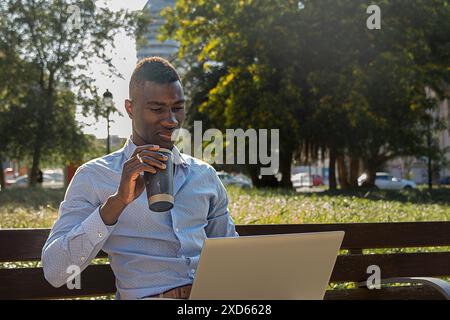 Afroamerikanischer Geschäftsmann, der im Freien mit Laptop arbeitet und Kaffee genießt. Business, Produktivität und Work-Life-Balance-Konzept in der Natur Stockfoto