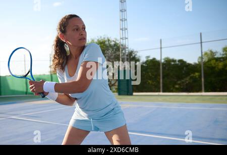 Eine Frau in sportlicher Kleidung, die Tennis auf einem Platz im Freien spielt. Sie sieht fokussiert und entschlossen aus, hält einen Tennisschläger bereit. Stockfoto