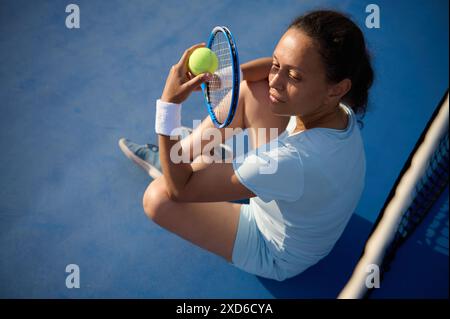 Eine Frau sitzt auf einem Tennisplatz mit Tennisschläger und -Ball und macht eine Pause vom Spielen. Sie sieht entspannt und zufrieden aus. Stockfoto