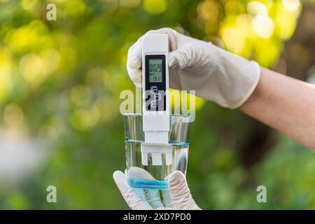 PH-Messgerät in der Hand mit Handschuhen, Glas Wasser auf verschwommenem Hintergrund der Natur. Messung der Eigenschaften von Trinkwasser. Wasserhärte. ve Stockfoto