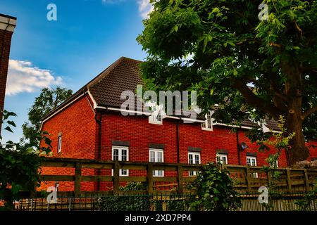 Ein rotes Backsteinhaus mit einem braun gekachelten Dach, umgeben von einem Holzzaun und üppigen grünen Bäumen. Der Himmel ist blau mit ein paar Wolken. Stockfoto