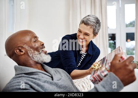 Überraschung, Sofa und aufregendes Paar mit Geschenk, um Geburtstag, Jubiläum oder Dankbarkeit zu Hause zu feiern. Liebe, reife Frau und Mann auf Couch mit Geschenk Stockfoto