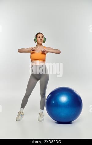 Eine schwangere Frau in Activewear steht neben einem hellblauen Trainingsball auf grauem Hintergrund. Stockfoto