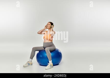 Eine junge, sportlich schwangere Frau in Aktivkleidung und Kopfhörern, die auf einem Turnball sitzt. Stockfoto