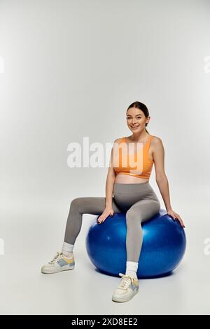 Eine junge, sportliche schwangere Frau in aktiver Kleidung sitzt anmutig auf einem hellblauen Trainingsball vor neutralem Hintergrund. Stockfoto