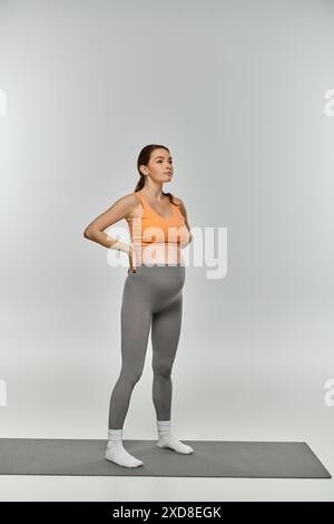 Eine sportliche schwangere Frau in Trainingskleidung steht selbstbewusst auf einer Matte mit Händen auf den Hüften und zeigt ihre Kraft und Vitalität. Stockfoto