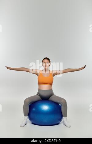 Eine junge, schwangere Frau in aktiver Kleidung balanciert sich anmutig auf einem leuchtend blauen Ball in einer grauen Umgebung. Stockfoto