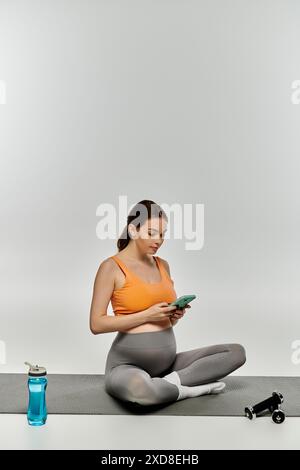 Sportliche schwangere Frau in Activewear, die auf Yogamatte sitzt und das Telefon überprüft. Stockfoto