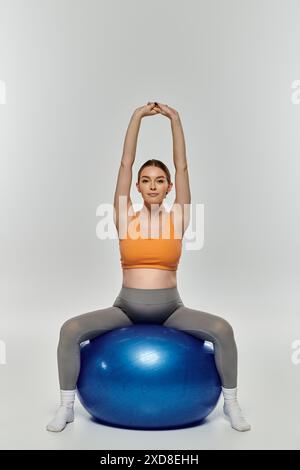 Eine schwangere Frau in Activewear balanciert auf einem blauen Trainingsball in einer Yoga-Pose vor grauem Hintergrund. Stockfoto
