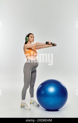 Sportliche schwangere Frau in aktiver Kleidung, die auf einem Trainingsball mit Kopfhörern auf grauem Hintergrund trainiert. Stockfoto