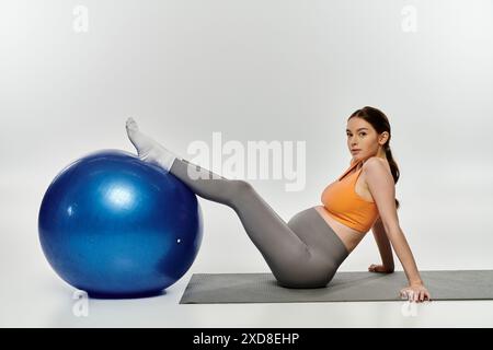 Eine sportlich schwangere Frau in aktiver Kleidung führt auf einem Trainingsball vor grauem Hintergrund Kernübungen durch. Stockfoto