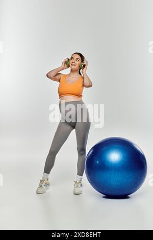 Eine sportlich schwangere Frau in Aktivkleidung, die neben einem hellblauen Trainingsball auf grauem Hintergrund steht. Stockfoto