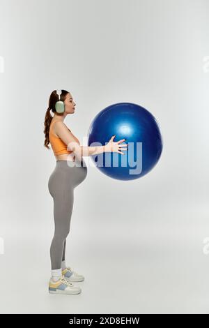 Eine junge, sportliche schwangere Frau in Aktivkleidung hält einen großen blauen Ball auf grauem Hintergrund. Stockfoto