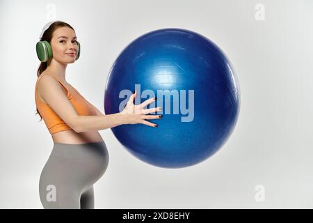 Eine junge schwangere Sportfrau in Aktivkleidung balanciert einen großen blauen Ball in ihren Händen auf grauem Hintergrund. Stockfoto