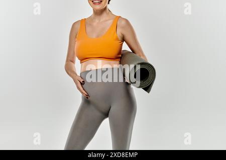 Eine junge, schwangere Frau in aktiver Kleidung lächelt, während sie eine Yogamatte hält. Stockfoto