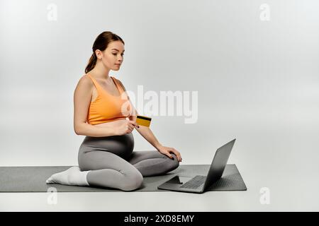Eine schwangere Frau in Aktivkleidung sitzt auf einer Yogamatte, während sie an einem Laptop arbeitet. Stockfoto