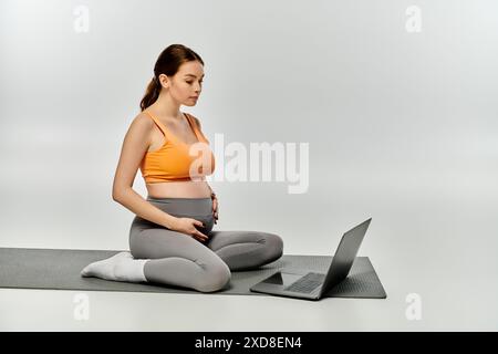 Eine sportliche schwangere Frau sitzt auf einer Yoga-Matte und Multitasking mit einem Laptop. Stockfoto