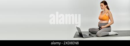 Eine schwangere Frau in Activewear sitzt ruhig auf einer Yogamatte, in einer friedlichen Umgebung mit einem Laptop. Stockfoto