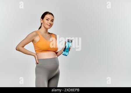Eine junge und sportliche schwangere Frau in aktiver Kleidung posiert selbstbewusst, während sie eine Wasserflasche auf grauem Hintergrund hält. Stockfoto