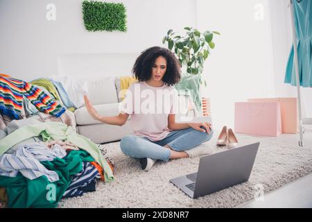 Foto eines bezaubernden hübschen Mädchens, das trendiges Outfit trägt und auf dem Boden sitzt und Schild ohne Sortimentskleidung zeigt Stockfoto