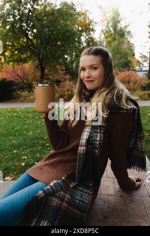 Frau in einem braunen Pullover, der einen braunen Becher hält, sitzt in einem Park Stockfoto