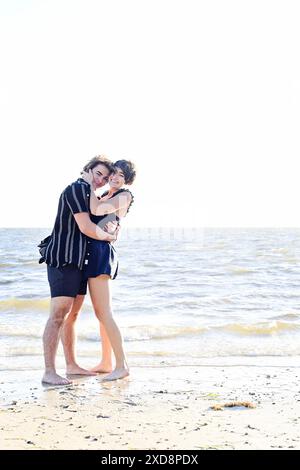 Zwei junge Leute umarmen sich am Strand, lächeln und genießen das Stockfoto