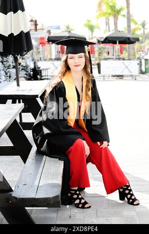 Weibliche Absolventin in Mütze und Kleid, sitzt draußen auf einer Bank, Klo Stockfoto