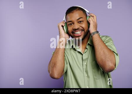 Ein glücklicher junger afroamerikanischer Mann mit Zahnspangen lächelt breit nach der Kamera, während er Kopfhörer auf einem leuchtend violetten Hintergrund trägt. Stockfoto