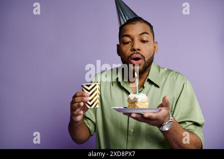 Ein junger afroamerikanischer Mann, der Kerze auf Geburtstagskuchen auf lila Hintergrund bläst. Stockfoto