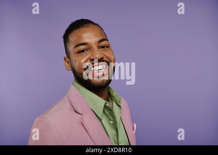 Junger afroamerikanischer Mann lächelt in einem stilvollen rosa Anzug und einem grünen Hemd vor einem leuchtend violetten Hintergrund. Stockfoto