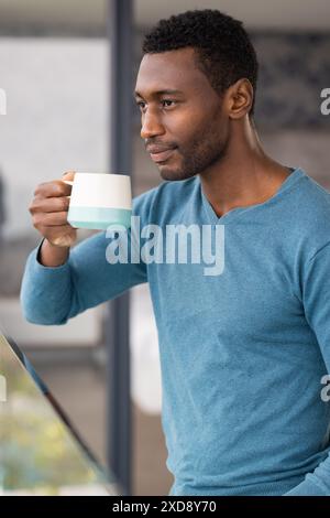 Einen Becher halten, ein Afroamerikaner in blauem Pullover, der aus dem Fenster blickt Stockfoto