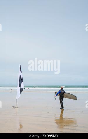 Ein einsamer männlicher Sufer mit seinem Surfbrett, der an einer schwarz-weiß karierten Flagge am Towan Beach in Newquay in Cornwall in Großbritannien vorbei geht. Stockfoto