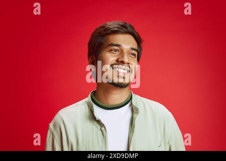 Porträt eines attraktiven, lächelnden jungen Mannes in lässiger Kleidung, der vor rotem Atelierhintergrund wegschaut. Stockfoto