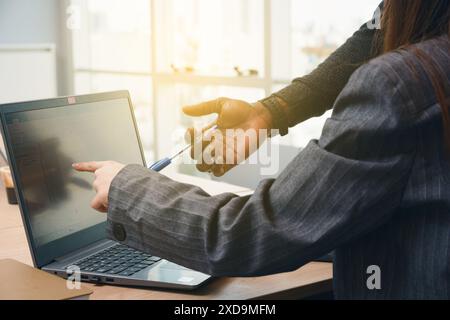 Ein Mann, der einen Stift hält, zeigt auf einen Laptop-Bildschirm mit einer Frau neben ihm. Die Frau trägt einen Anzug und der Mann trägt ihn. Der Laptop ist offen und Stockfoto