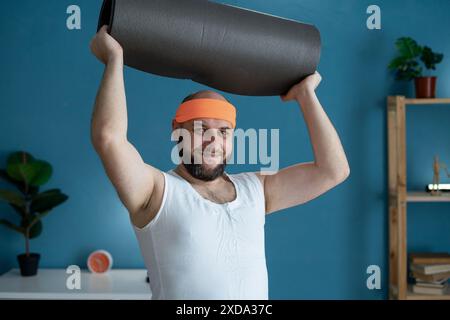 Mann mit Yogamatte bereit für das Training im Heimtrainer, mit Stirnband und athletischem Outfit Stockfoto