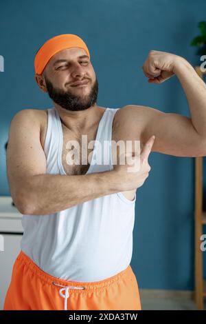 Porträt eines glücklichen Mannes, der seinen Bizeps beugt und während eines Workouts zu Hause selbstbewusst lächelt. Er trägt ein orangefarbenes Stirnband und Shorts. Persönliche Fitness Stockfoto